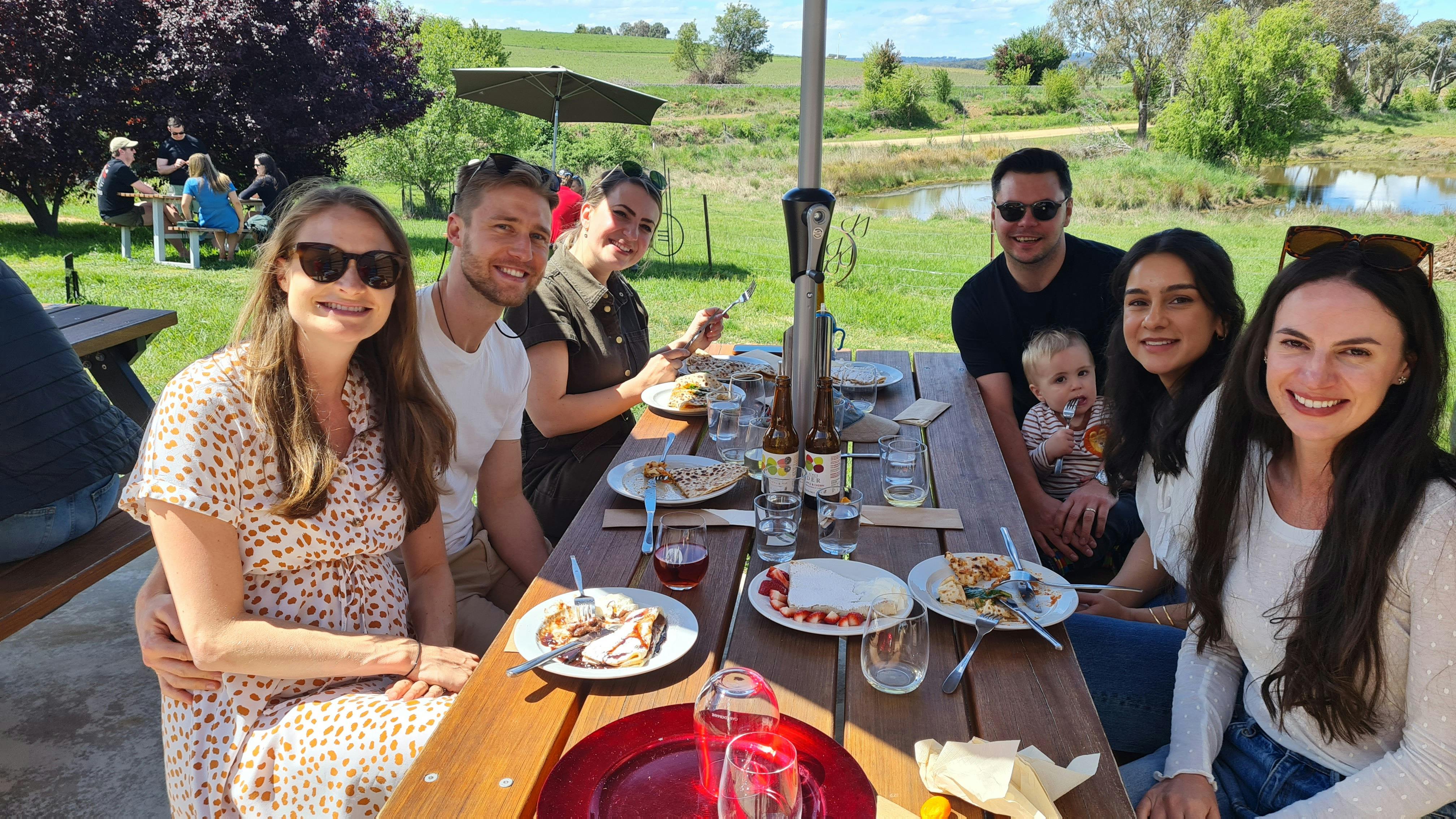 A group of young adults are seated under an umbrella at a picnic table with Crepes and cider