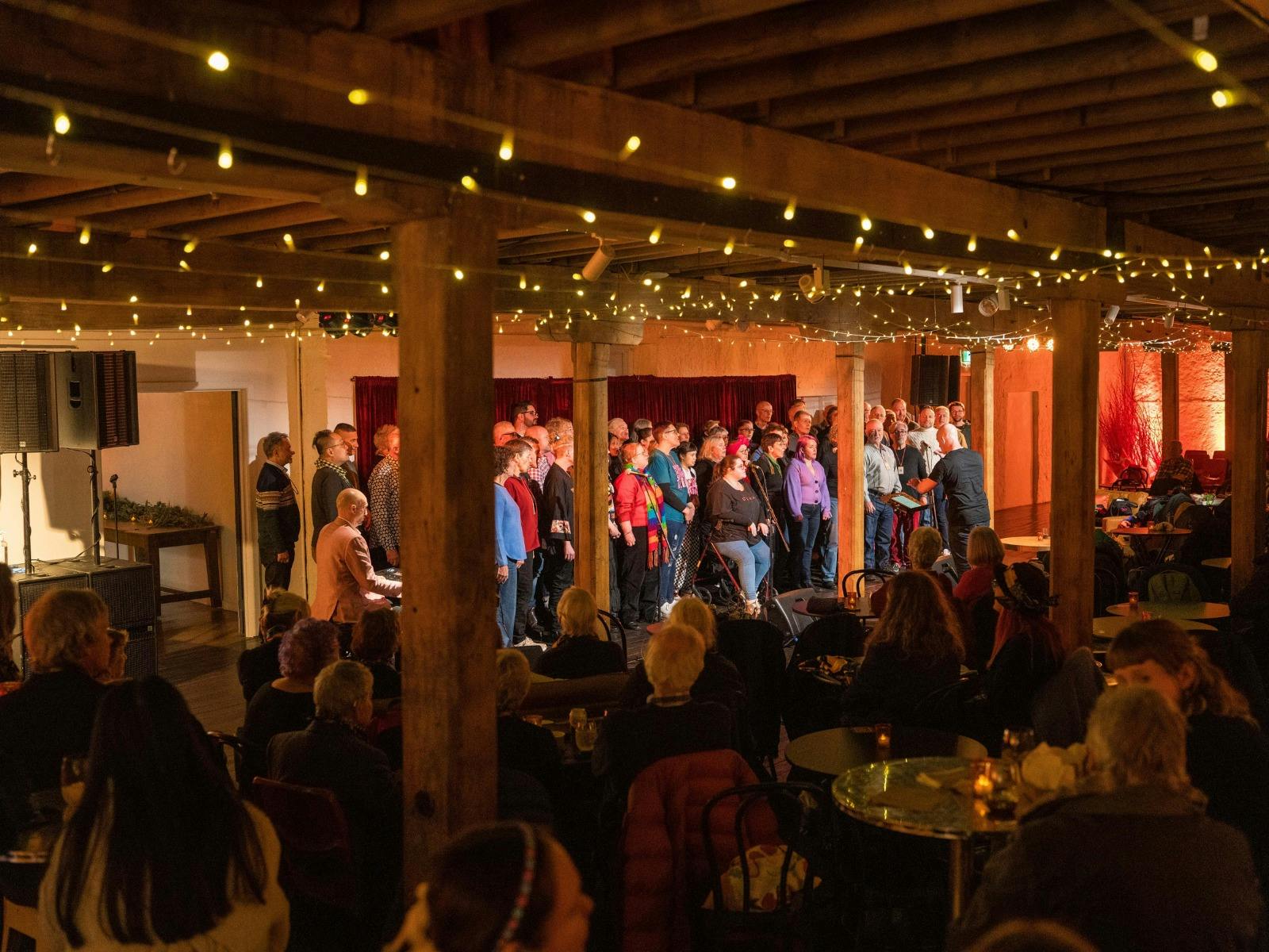Choir performing for a seated audience in a cozy room with wood beams and lights.