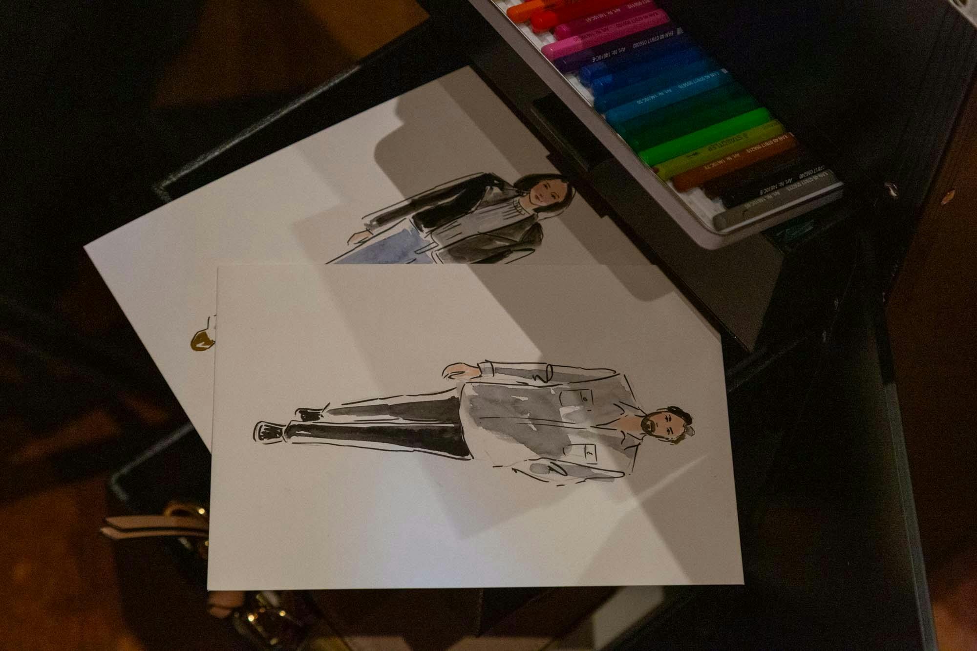 Two water colour fashion portraits of a man and a woman sitting on a table next to paints.