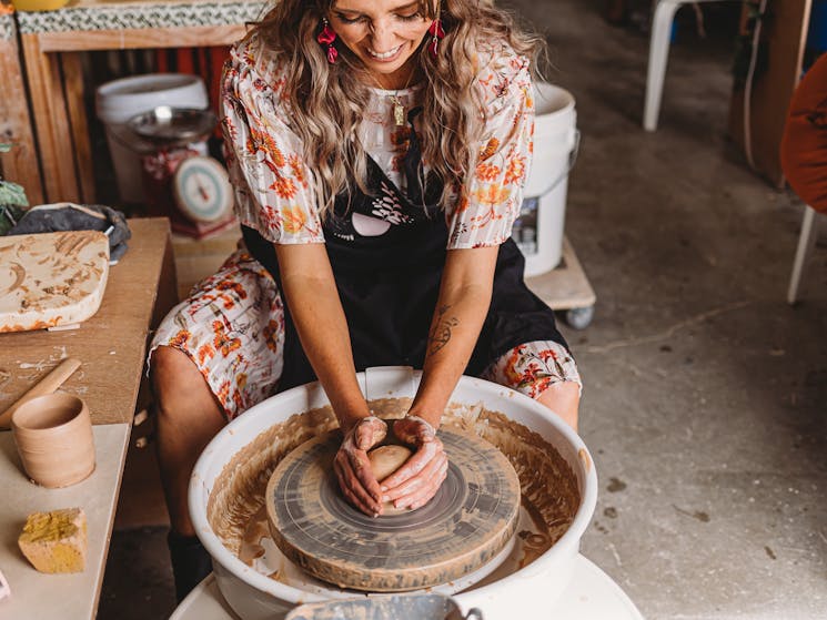 Photo shows female sitting at pottery wheel with hands on a ball of clay