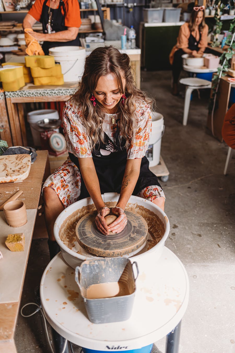 Photo shows female sitting at pottery wheel with hands on a ball of clay