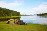 Tent and car are set on the lakes shores with pine forest in background.