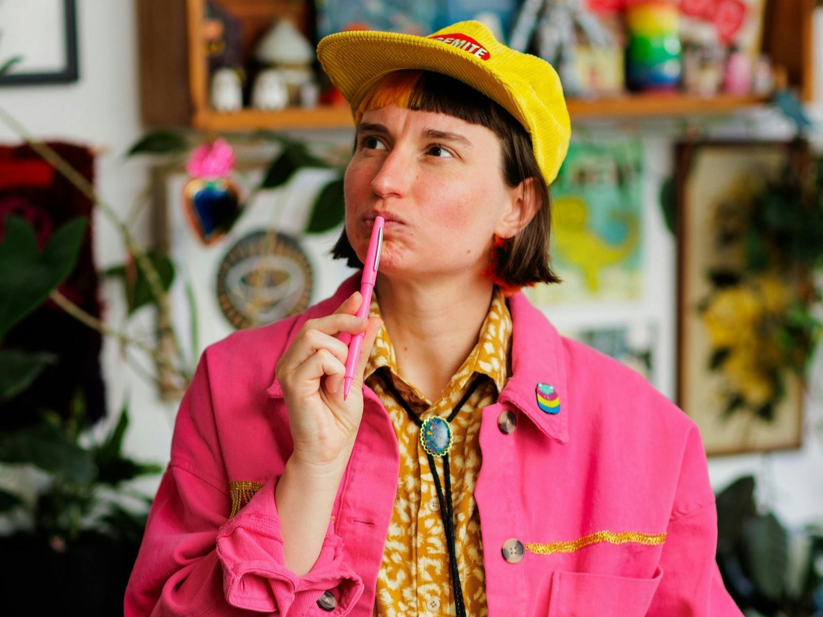 A person with short dark hair and a yellow cap looks thoughtfully upward while holding a pink pen.