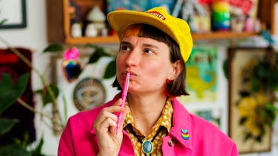 A person with short dark hair and a yellow cap looks thoughtfully upward while holding a pink pen.