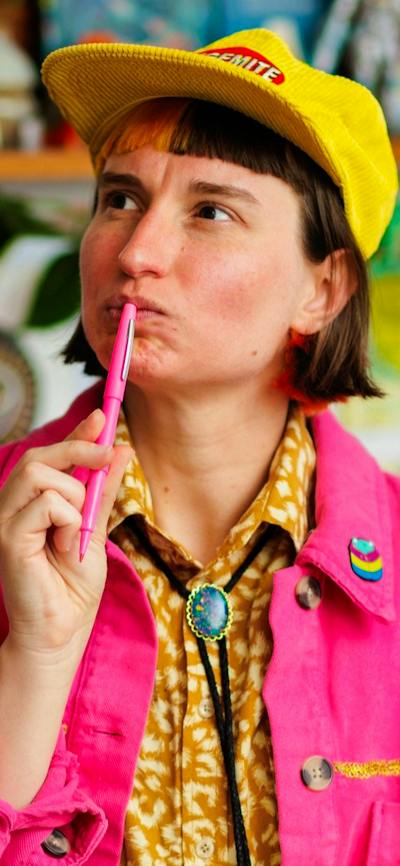A person with short dark hair and a yellow cap looks thoughtfully upward while holding a pink pen.