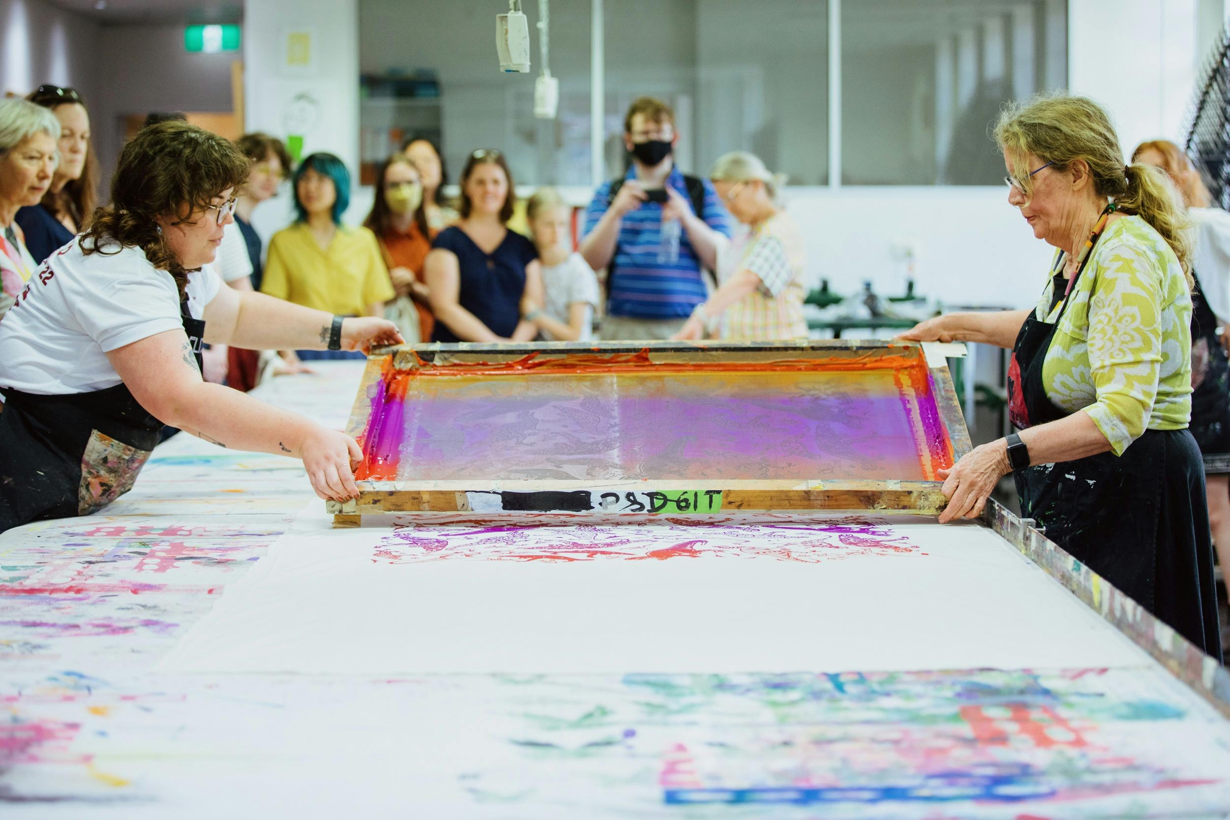 Two women lift a large colourful screen after printing