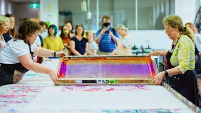 Two women lift a large colourful screen after printing