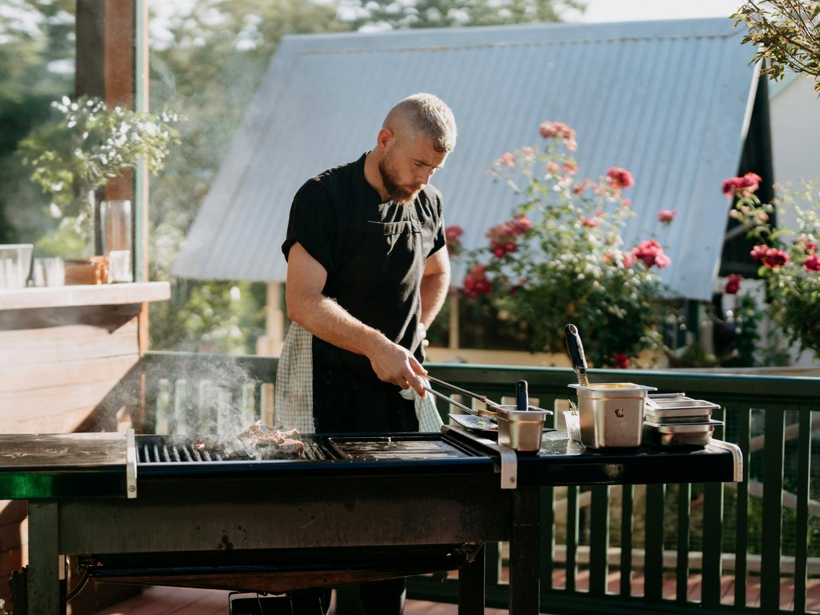 Chef standing at a barbeque on a sunlit deck, holding tongs cooking. roses bloom in garden nearby.
