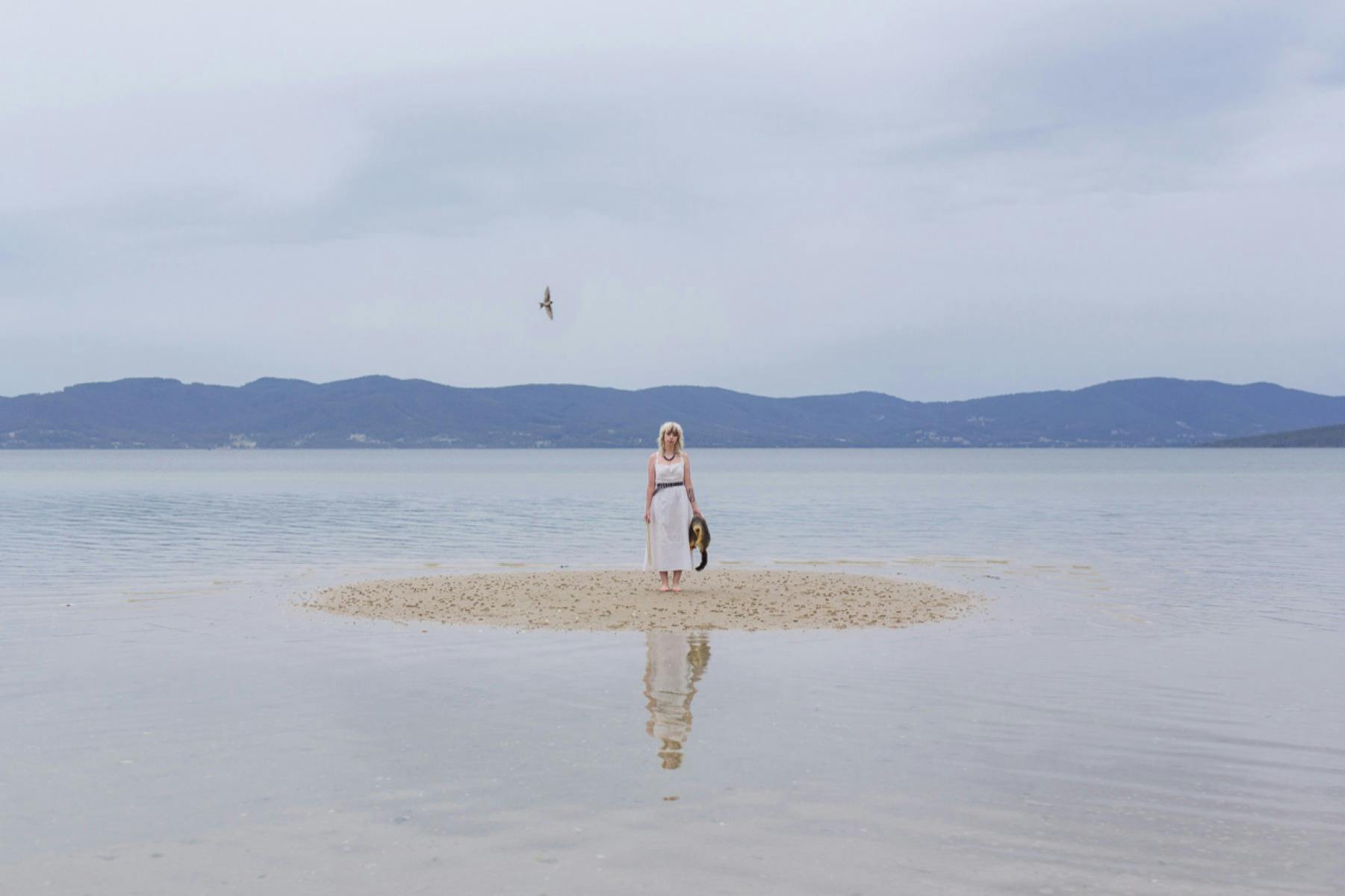 Image of a woman clothed in white on a sand mound surrounded by water. A bird flies overhead.