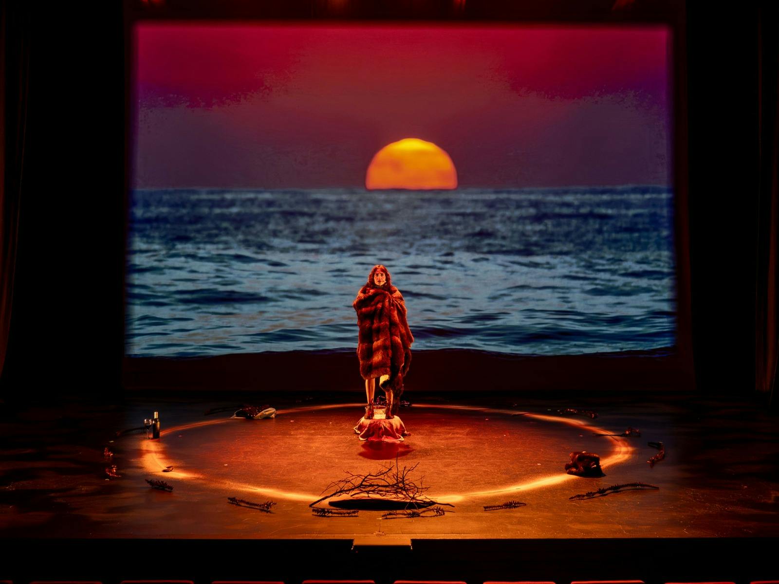 A person draped in a blanket stands center stage in front of a projected backdrop showing the ocean