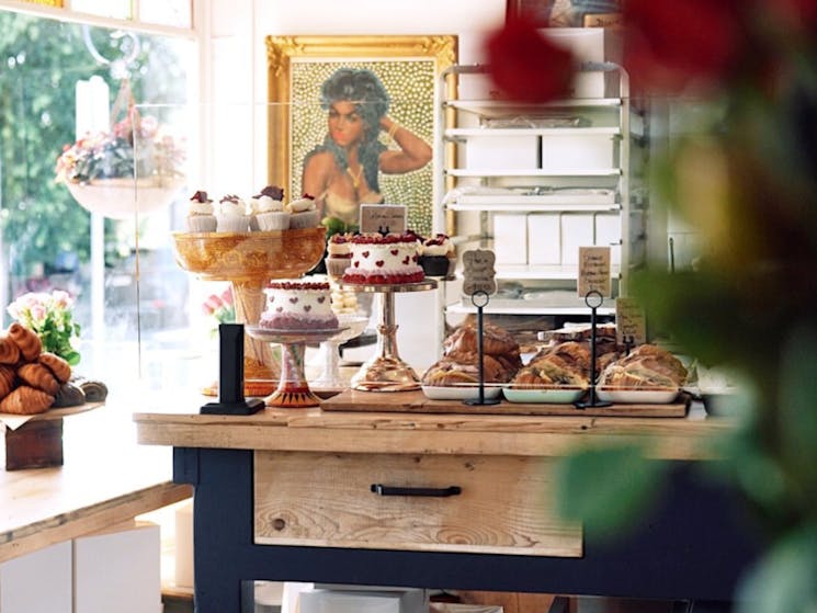 The inside of a shop with fresh cupcakes on a stand, croissants and flowers