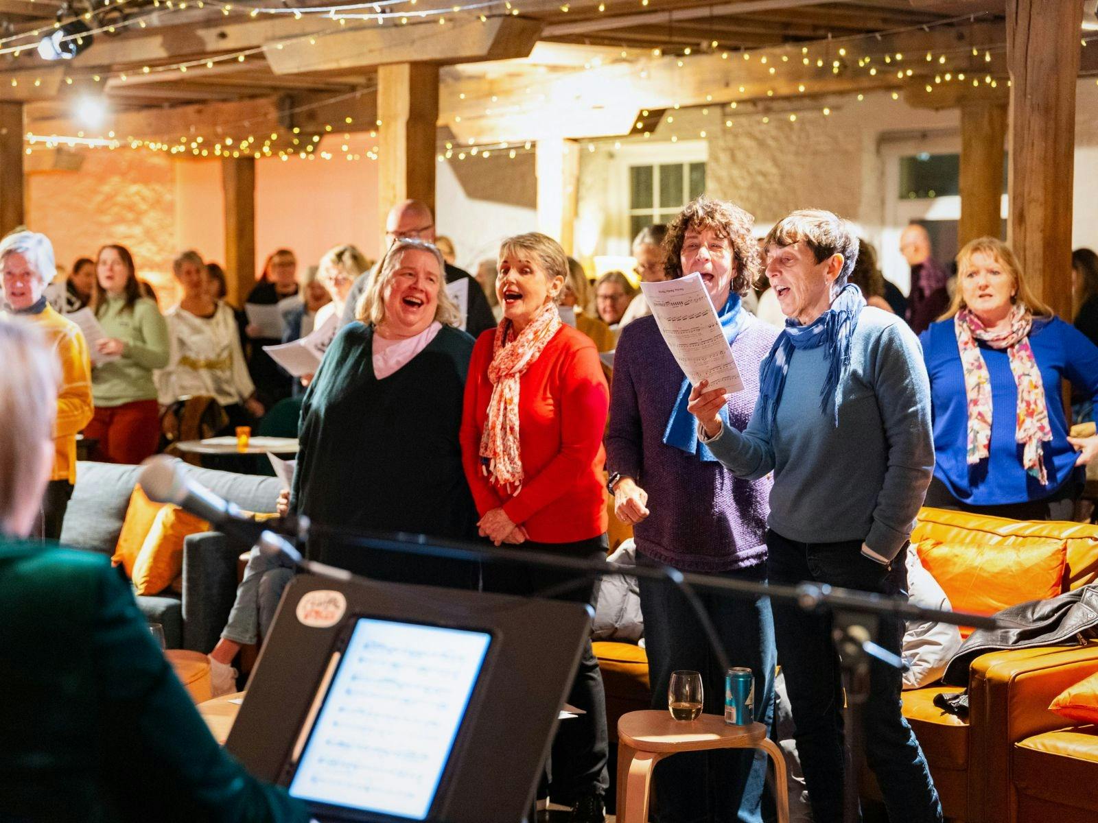 Four adults singing, holding sheet music, in a warmly lit, rustic interior.
