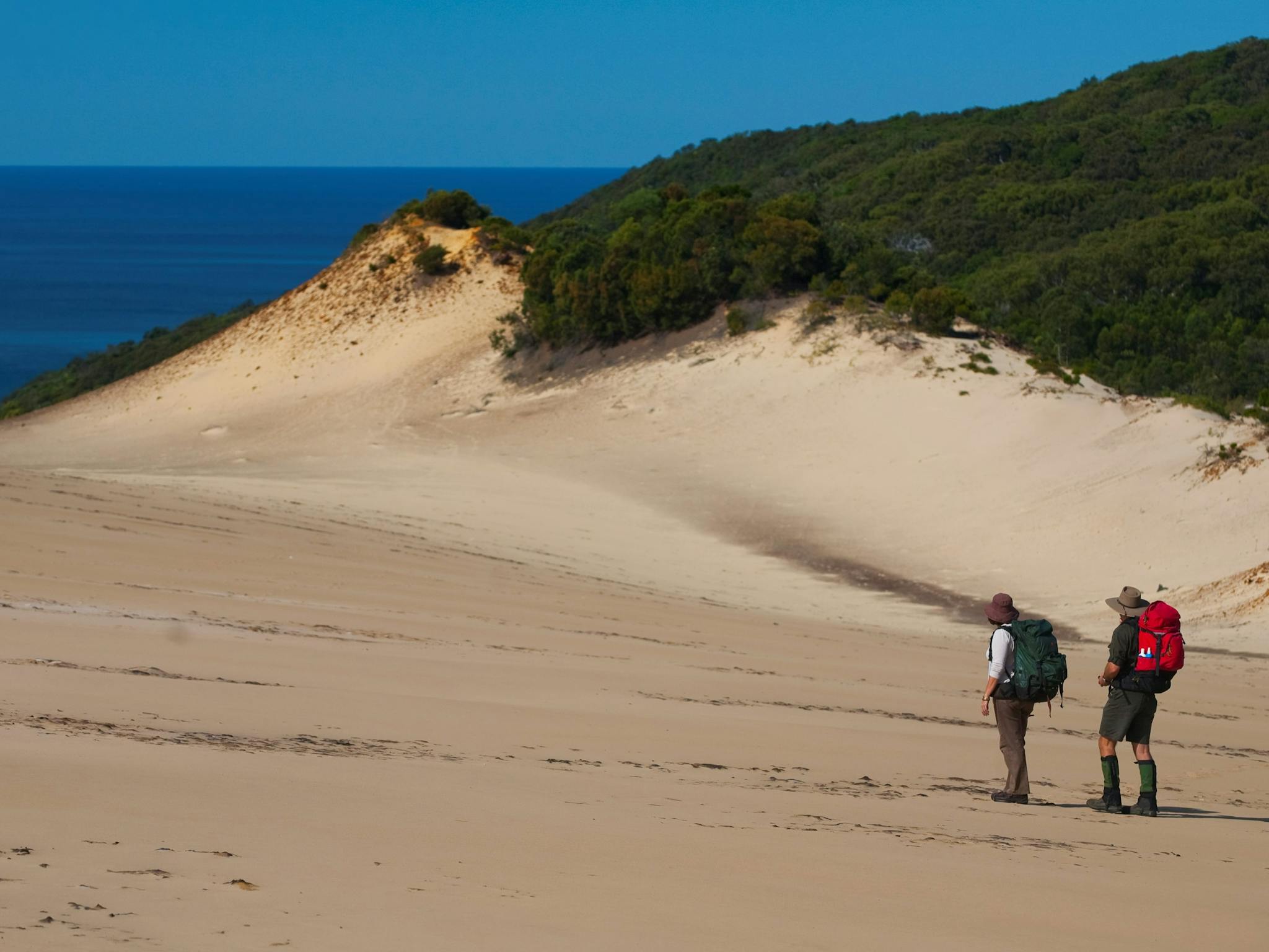 Two people with backpacks crossing a sandpatch with the ocean in the background. Two people with backpacks crossing a sandpatch with the ocean in the background.