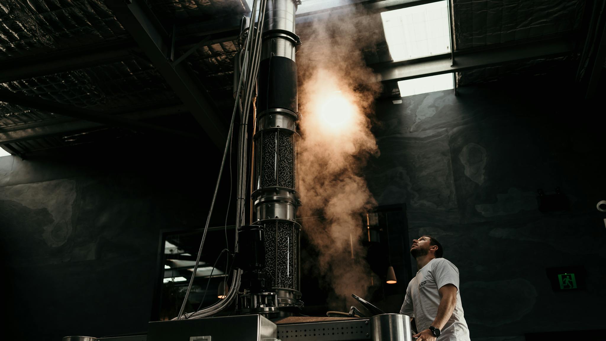 A man climbs a latter at a large stainless steel still and looks up as steam emerges