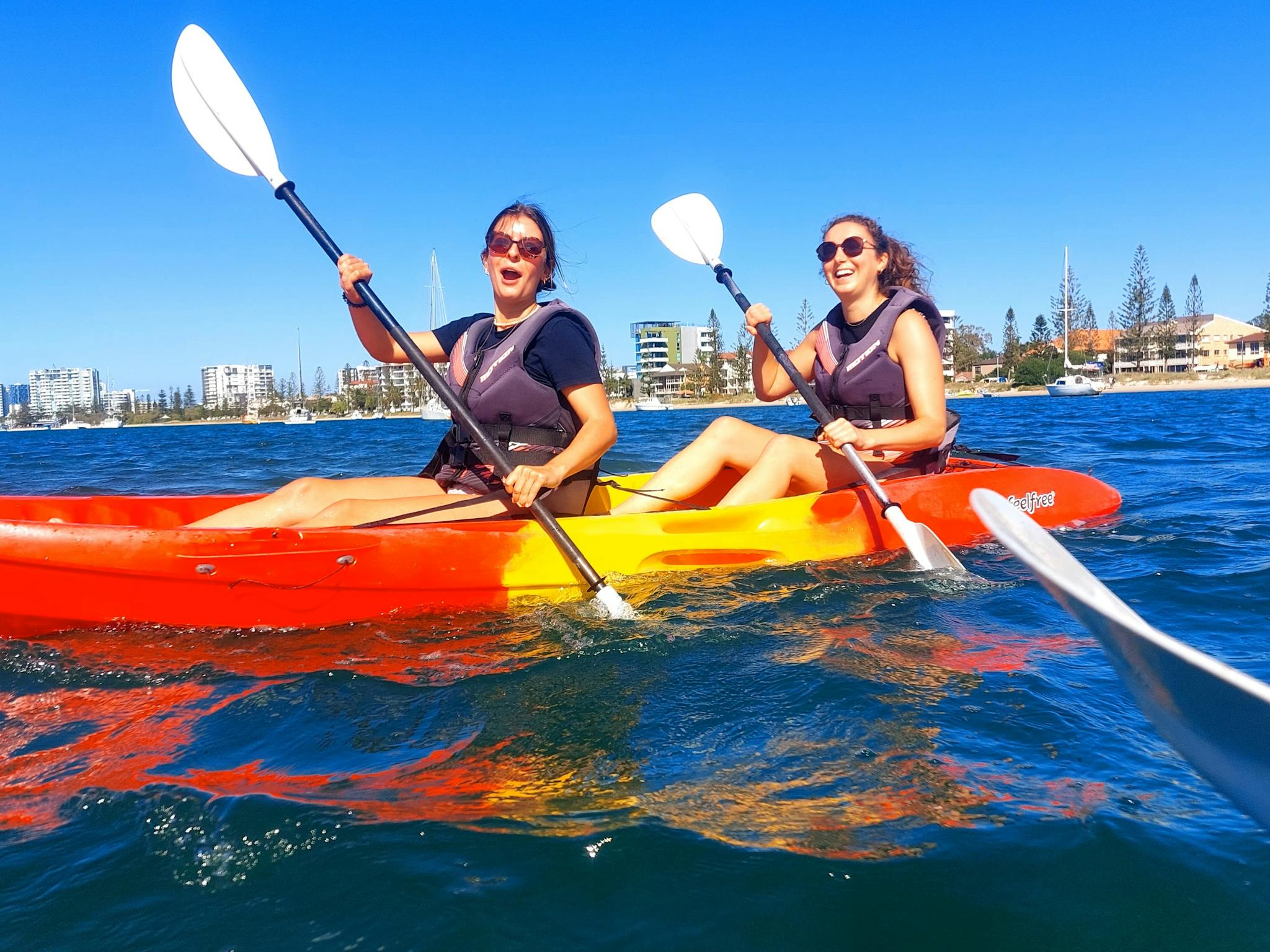wo friends paddle a vibrant kayak, smiling under clear skies with the Gold Coast skyline behind them wo friends paddle a vibrant kayak, smiling under clear skies with the Gold Coast skyline behind them