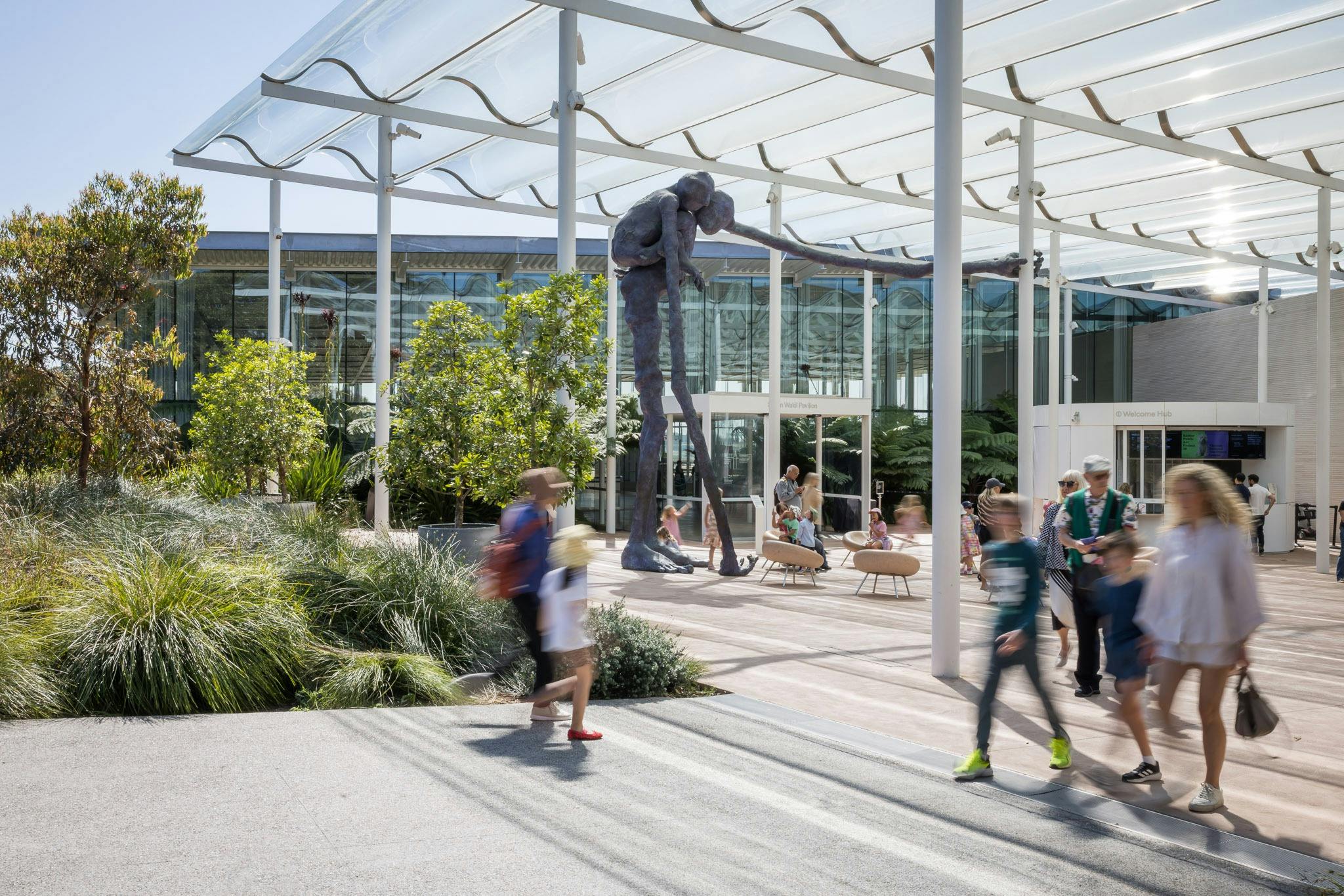 people arriving and leaving a glass welcome plaza at a gallery with a blue sculpture in the back