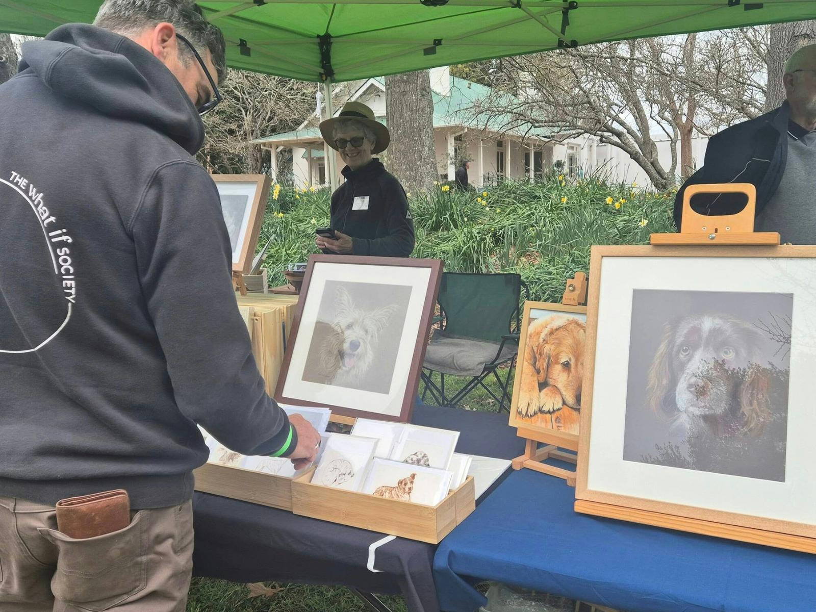 Stallholder displays their art. Image of dogs framed. Man in hoodie browses the display.