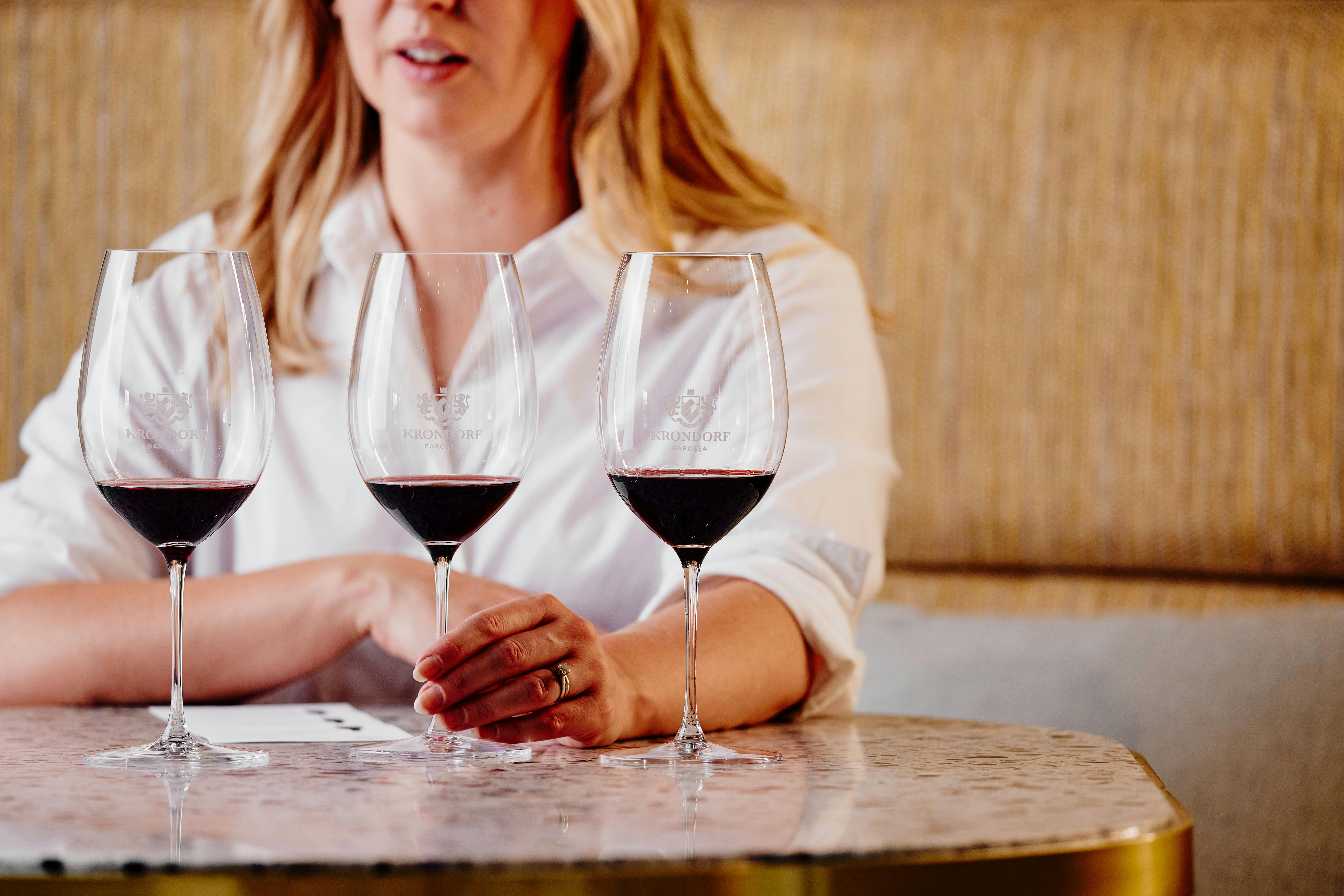 Three wine glasses with red wine sit on a stone table in front of a blonde woman in a white shirt