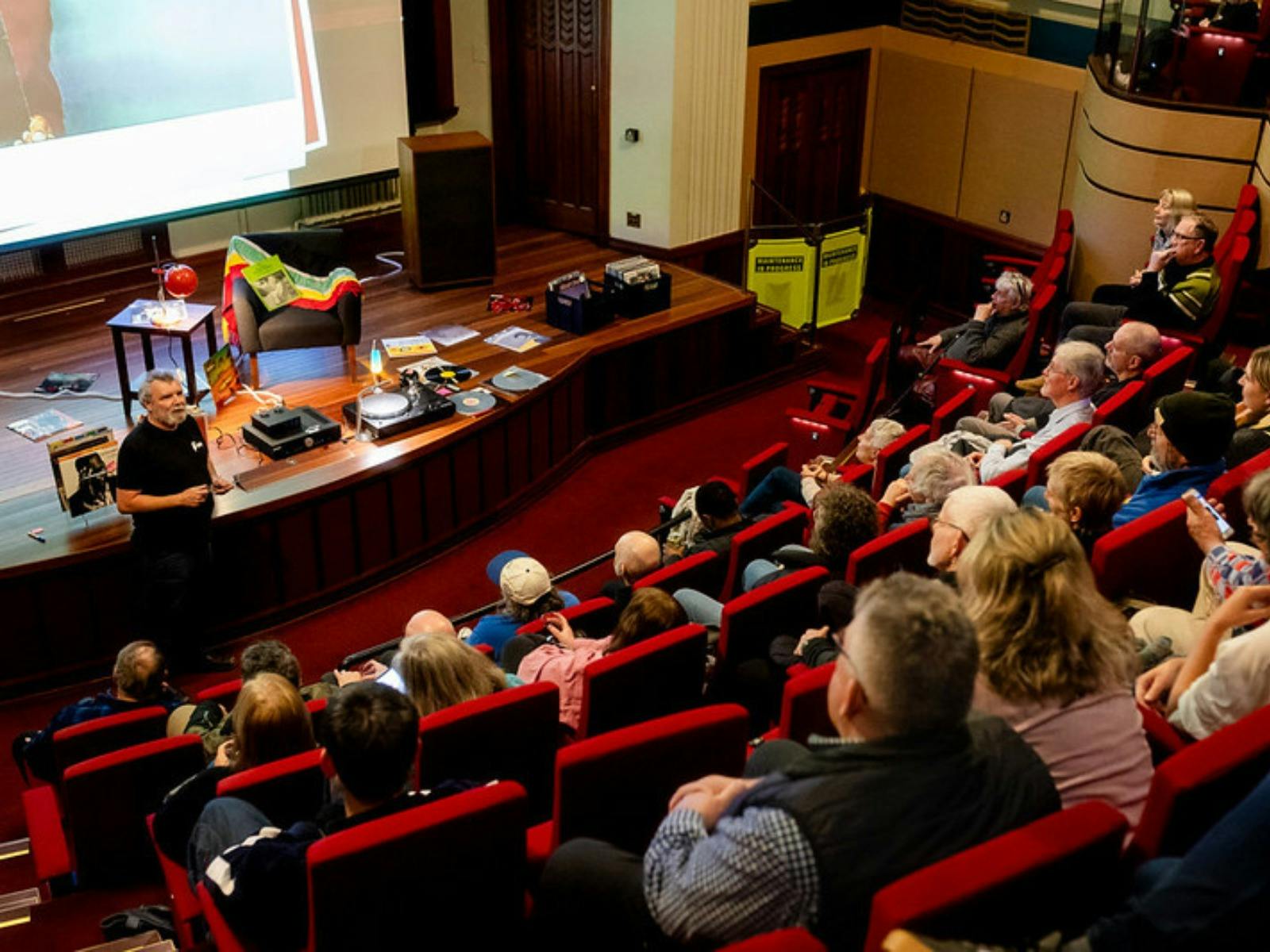 Audience in a theatre and a man at the front preparing to play a vinyl