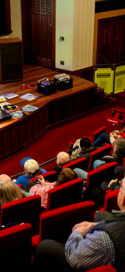 Audience in a theatre and a man at the front preparing to play a vinyl