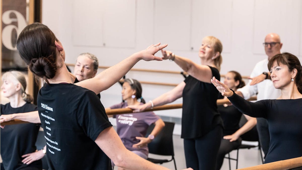 Ballet class for adults, some standing at the barre, and others seated focusing on arm positioning
