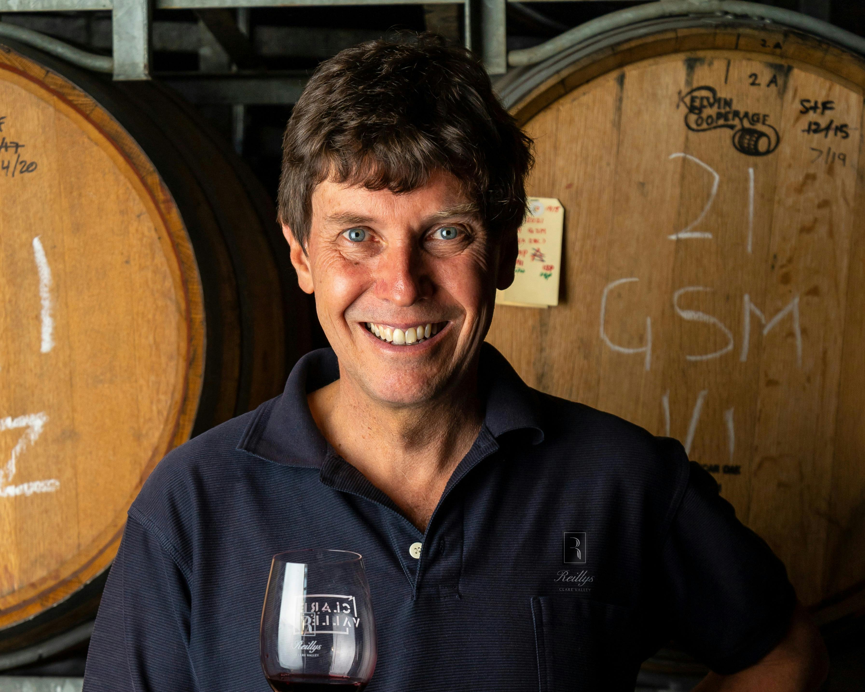Man with brown hair smiling at camera holding a glass of wine standing in front of wine barrels