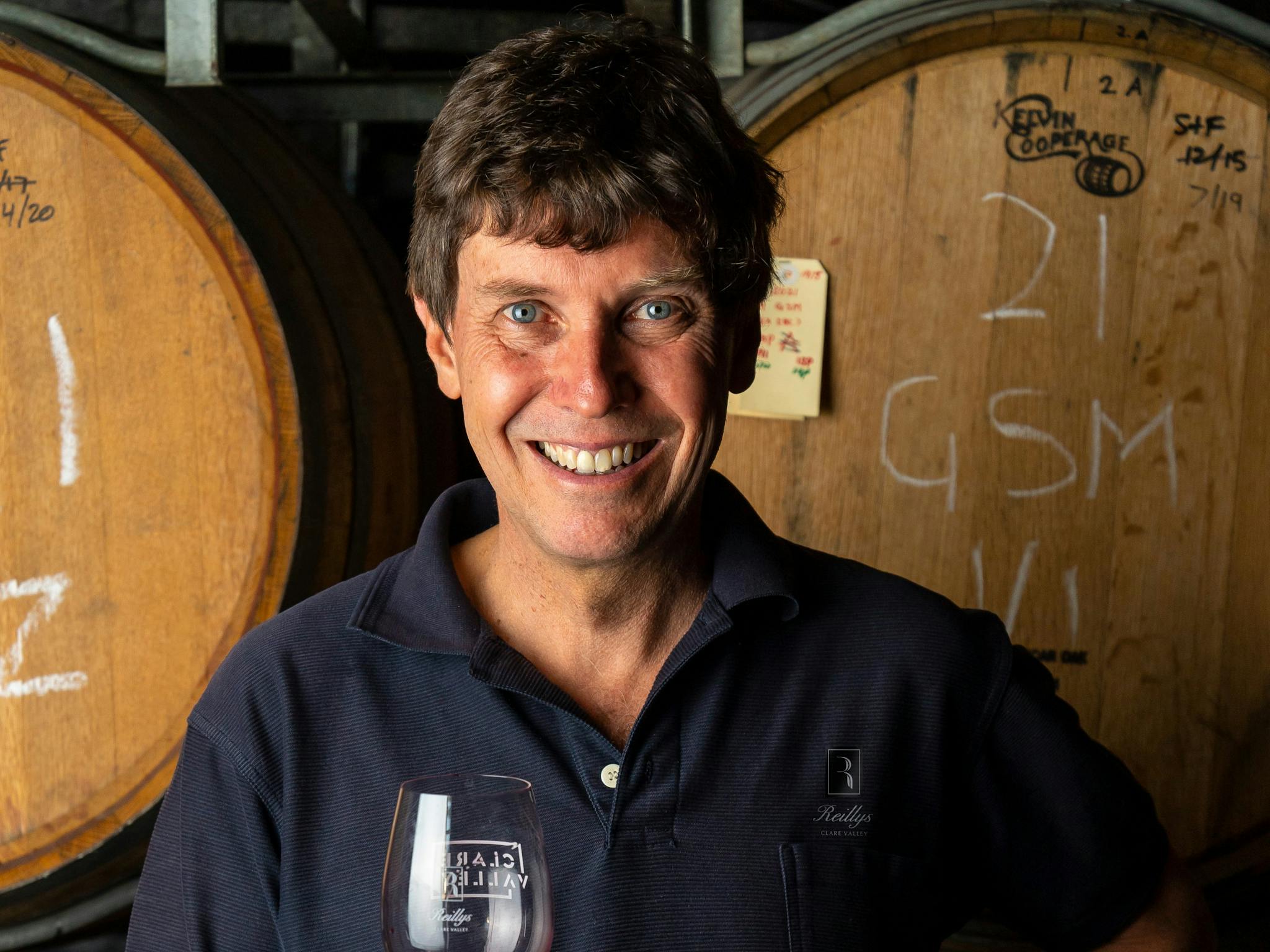 Man with brown hair smiling at camera holding a glass of wine standing in front of wine barrels