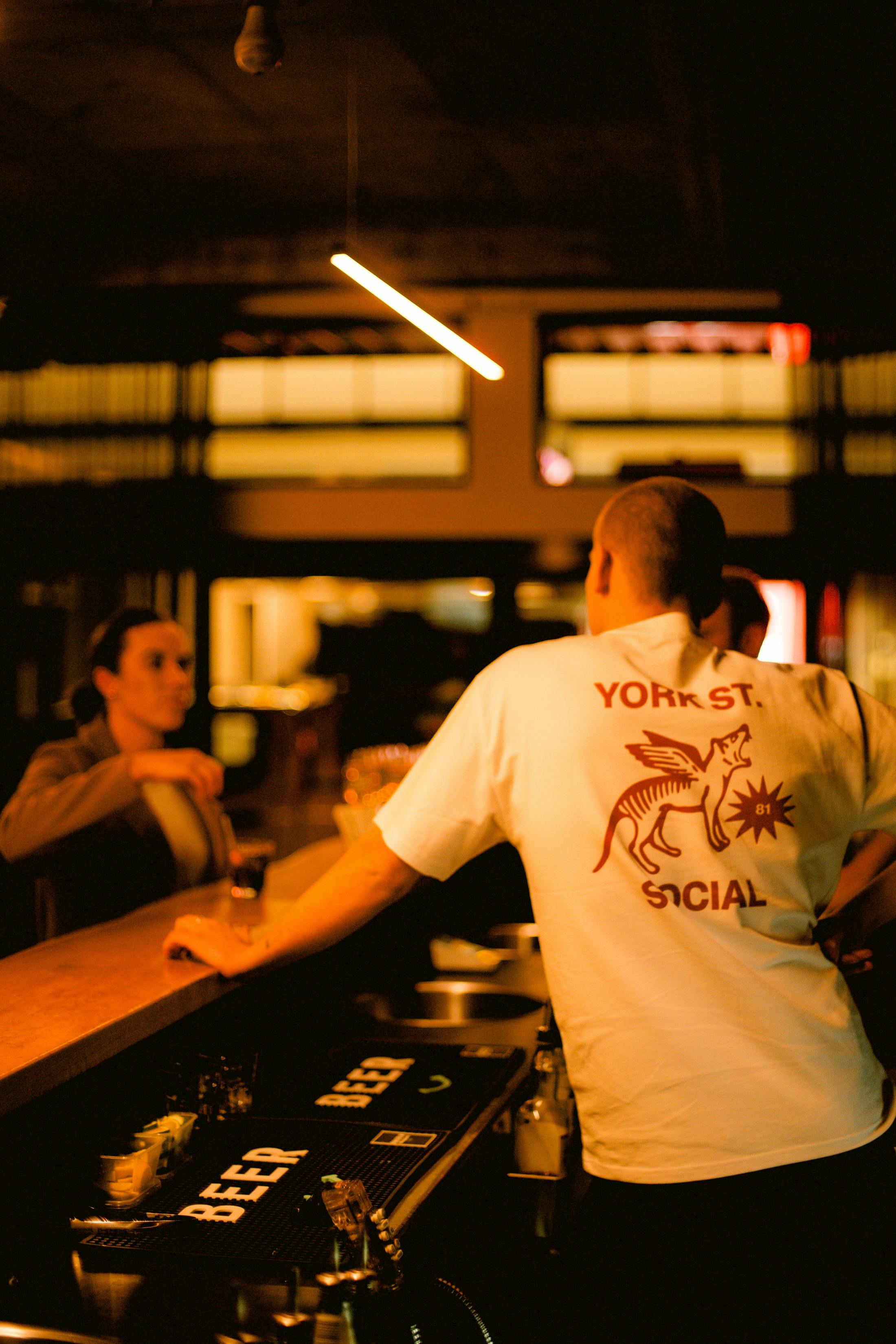 Man in white “York St. Social” shirt leaning at a bar, speaking with a woman holding a drink.