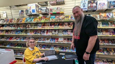 A woman sits at a table with signed books and pens, a man standing waits. Both smiling at the camera