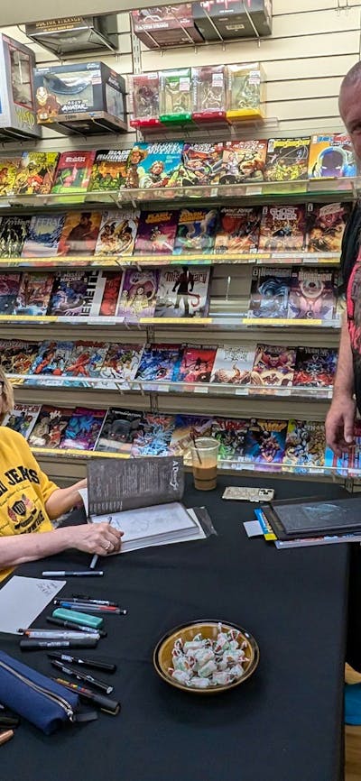 A woman sits at a table with signed books and pens, a man standing waits. Both smiling at the camera