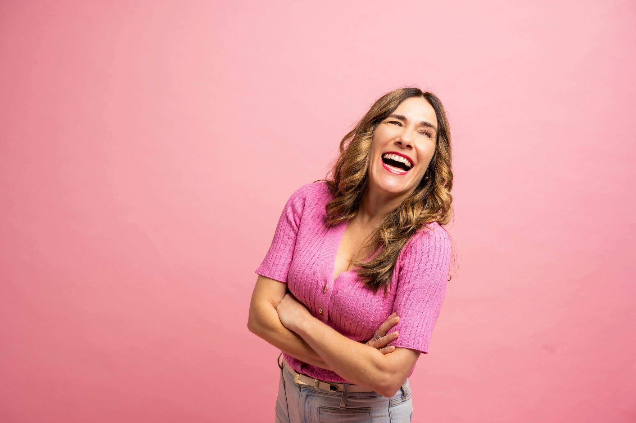 Marilyn is wearing a pink top with gold buttons and jeans, laughing with her arms crossed