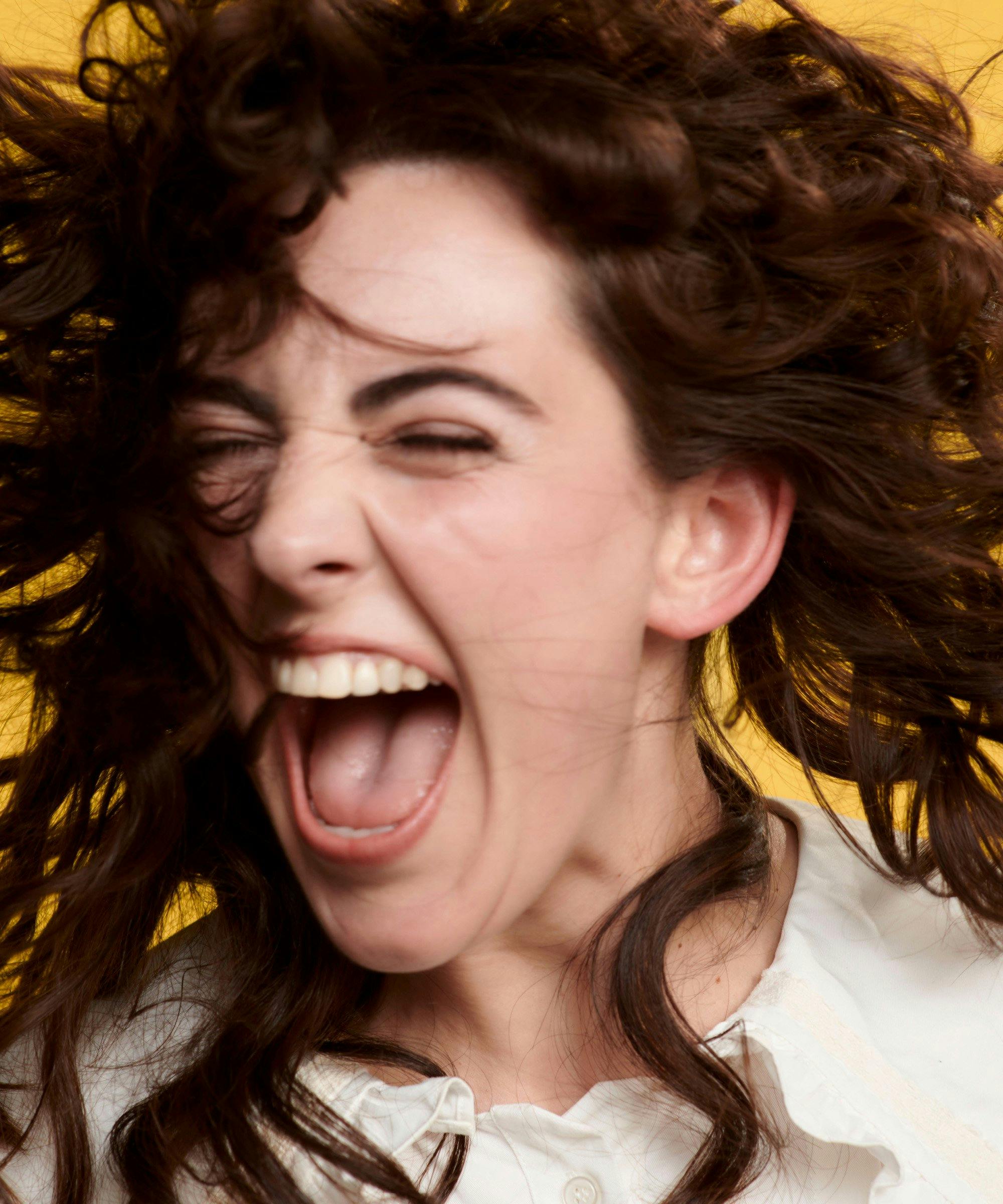 A girl with brown hair shakes her head while laughing against a yellow background.
