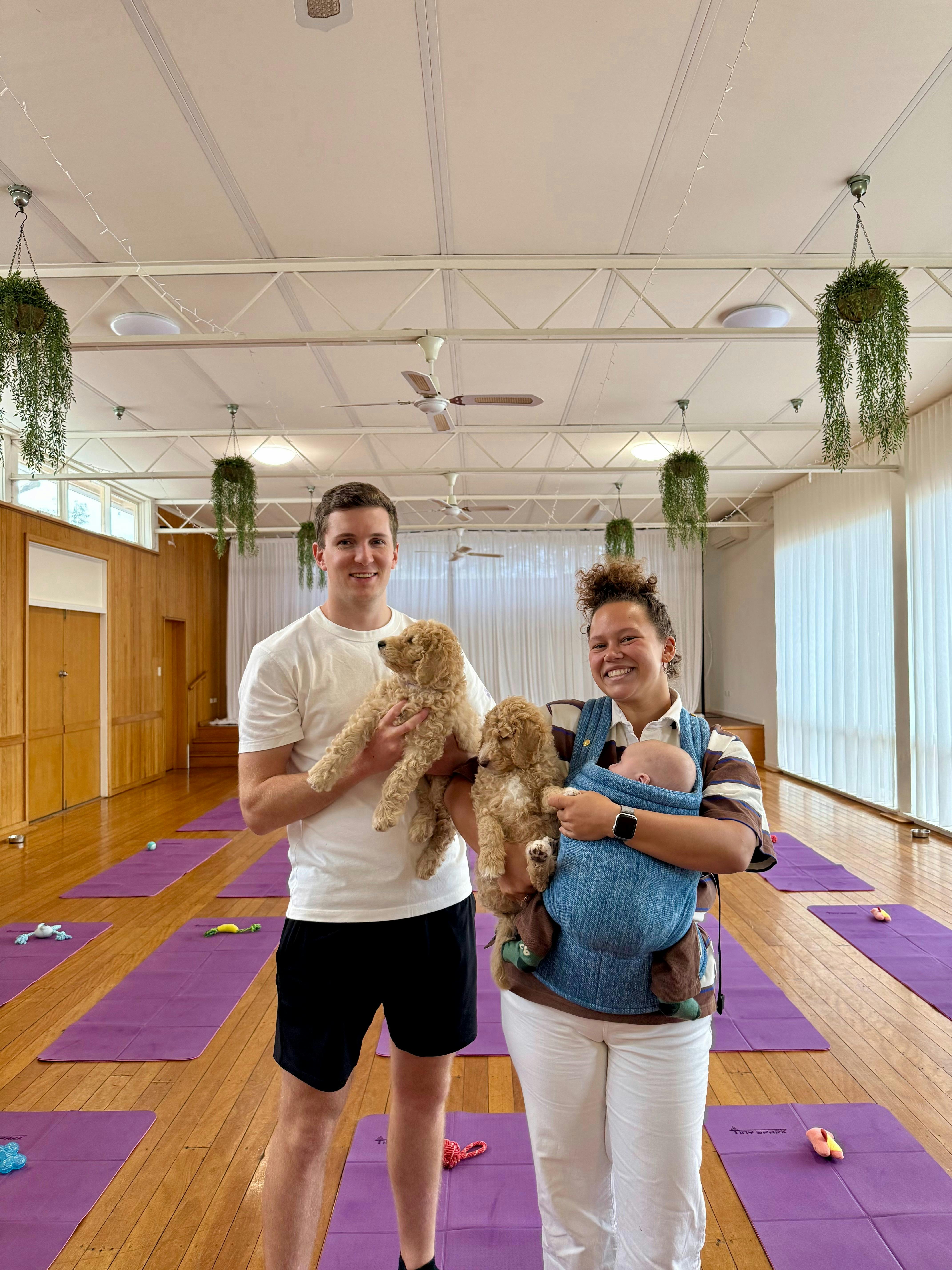 Two smiling participants hold puppies in a Canberra studio with purple yoga mats.