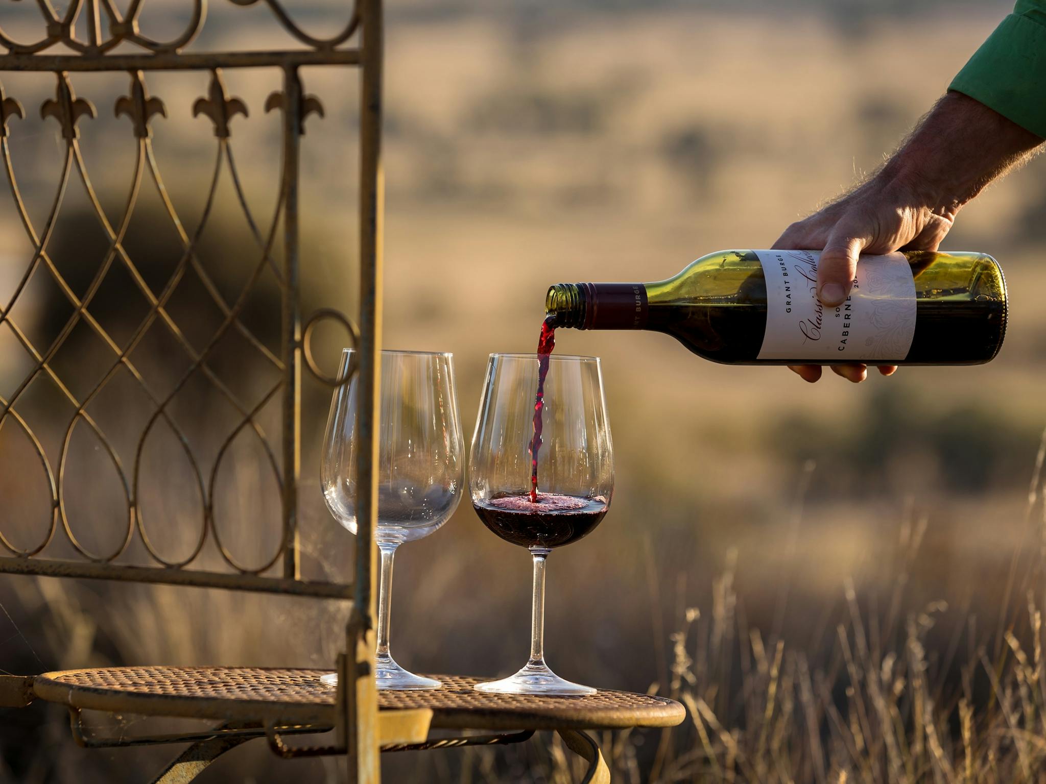 Outdoors, rural view; wine glasses perched on metal chair; hand pouring red wine