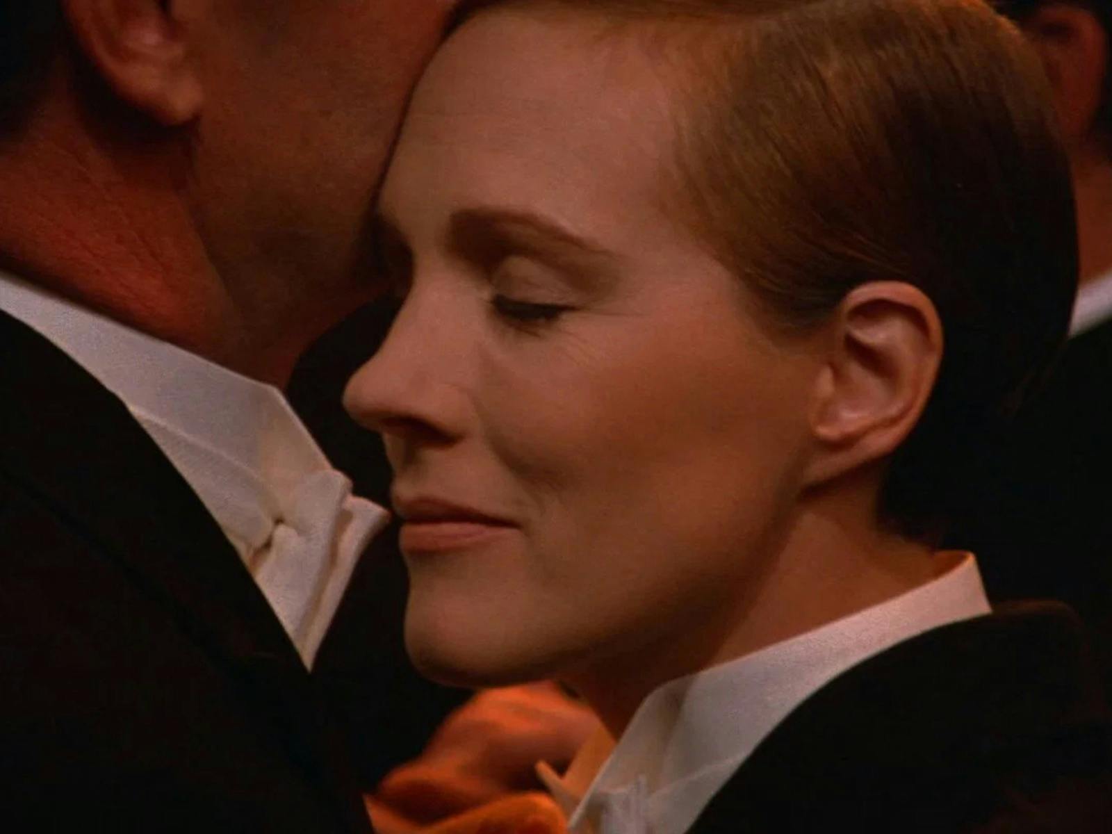 Close up of woman dressed in tuxedo with eyes closed pressed against chin of another person