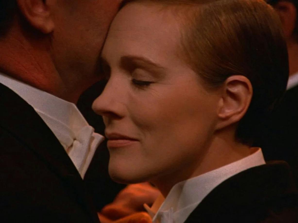 Close up of woman dressed in tuxedo with eyes closed pressed against chin of another person