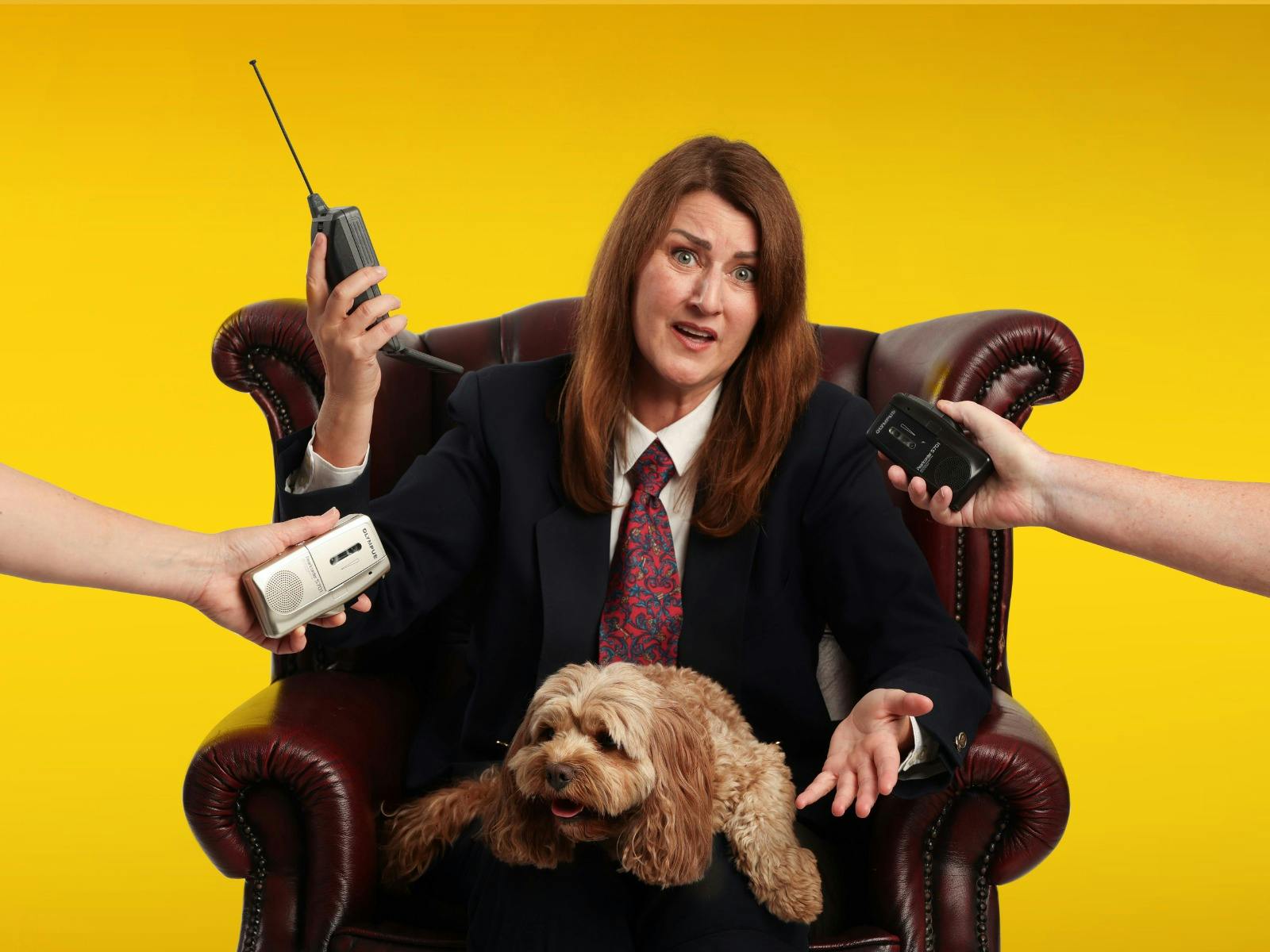 A woman sits in a brown leather armchair against a yellow backdrop with a dog and hands with items