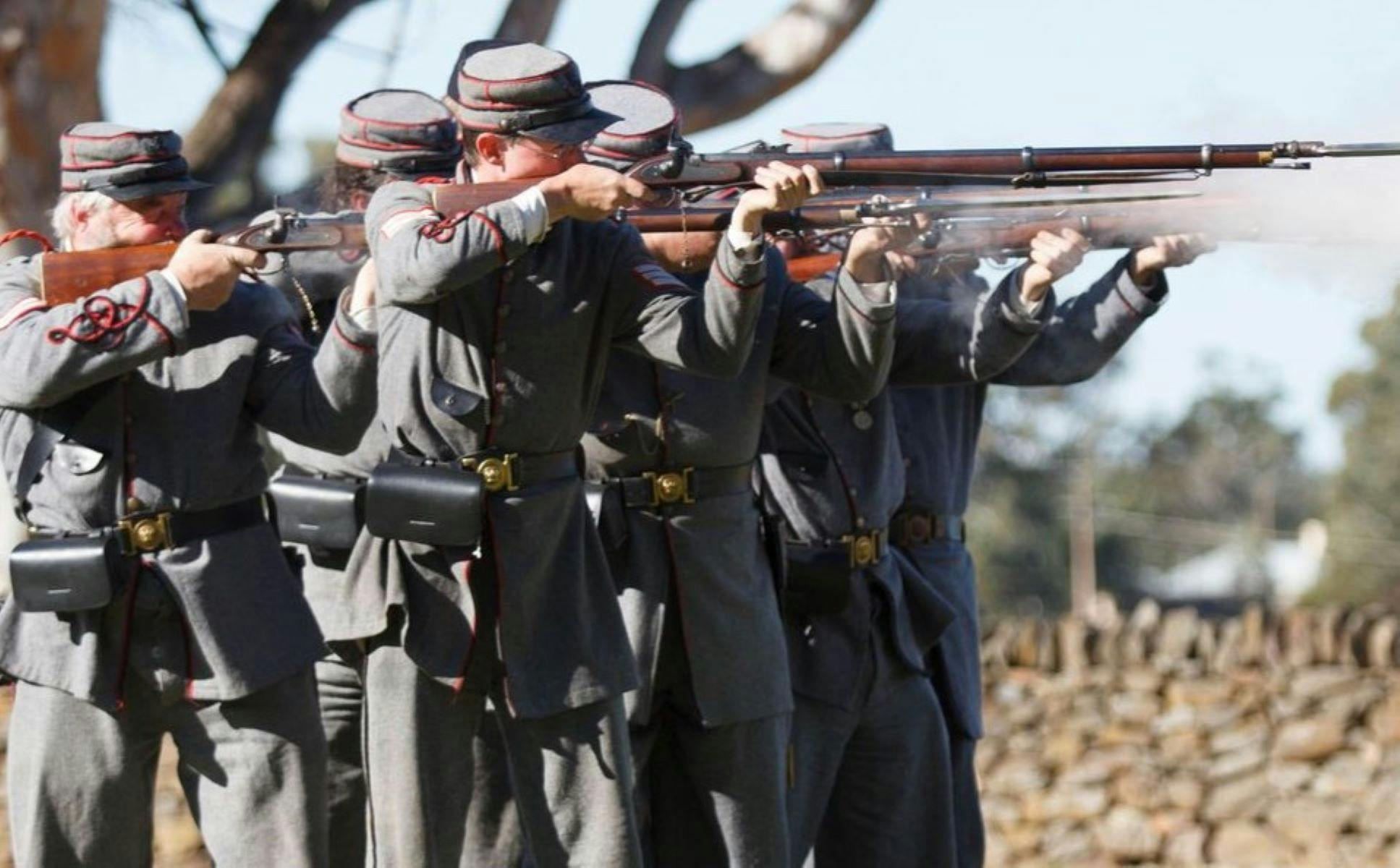 Group of people dressed in 1860 soldier uniforms, firing their muskets .