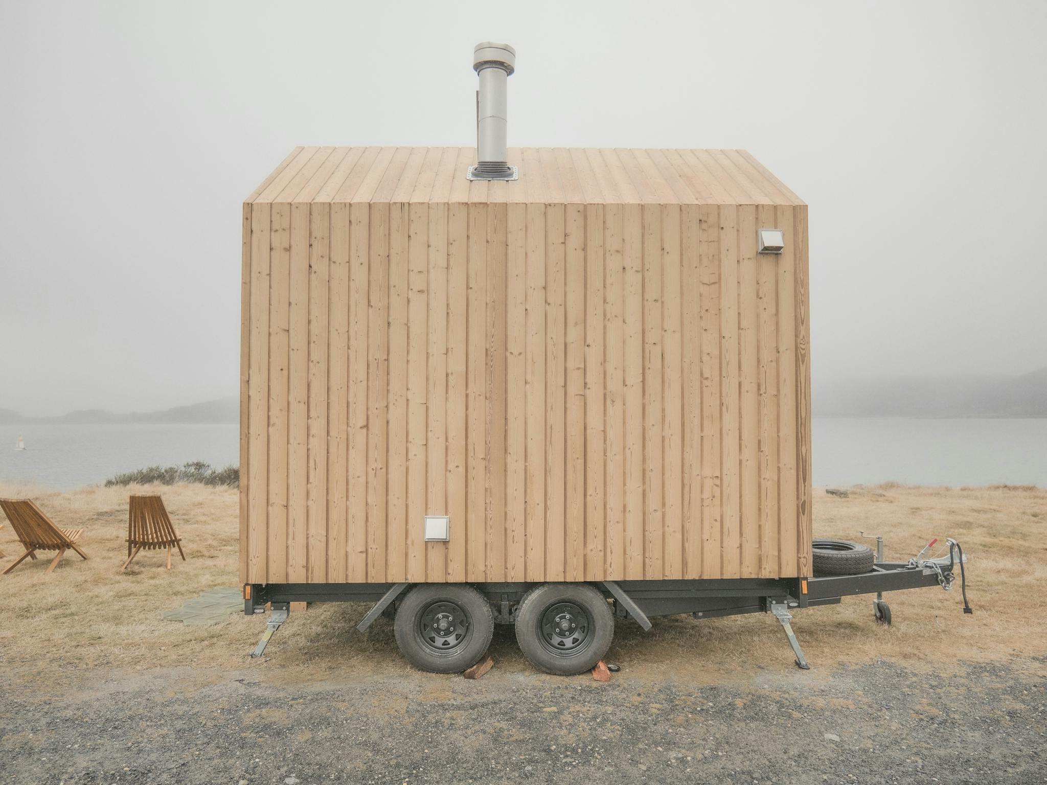 Light wood coloured mobile nature sauna parked in front of  a lake view on a foggy day