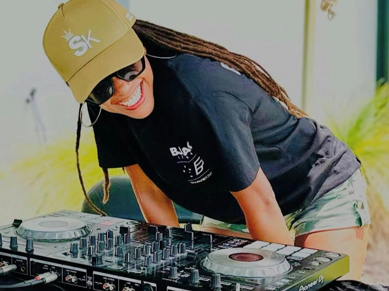 DJ grinning as she bends over desk wearing yellow hat and sunnies