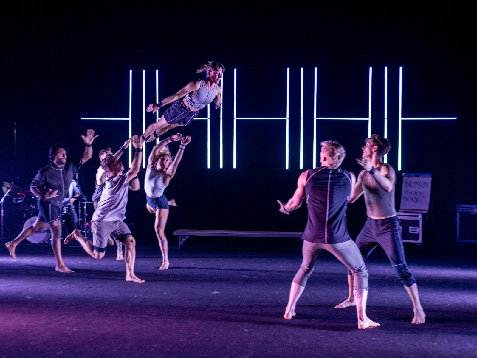 A group of acrobats performing stunts lit by coloured lighting with a neon clock in the background.
