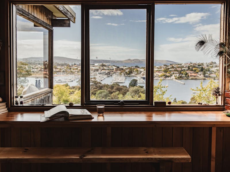 A book lays open next to a glass of water on a bench in front of wide windows showing the harbour.