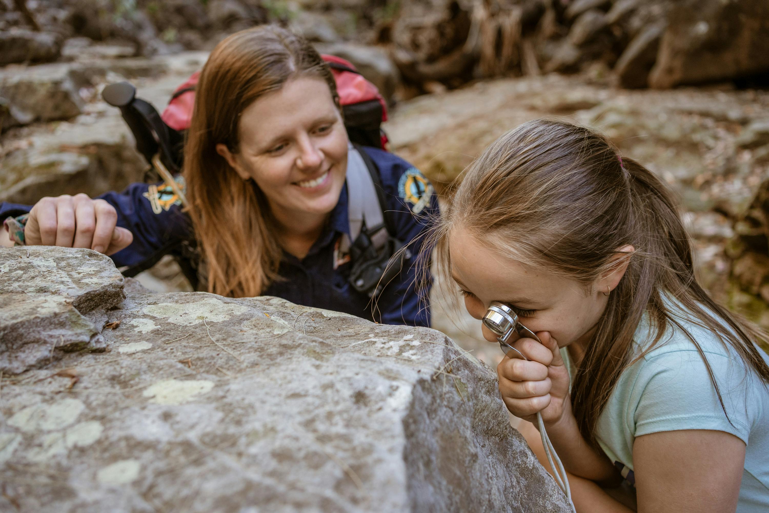 A tour guide assists a young girl as she peers through a hand lens at a tiny object, Carnarvon Gorge