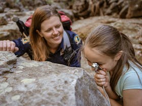 A tour guide assists a young girl as she peers through a hand lens at a tiny object, Carnarvon Gorge