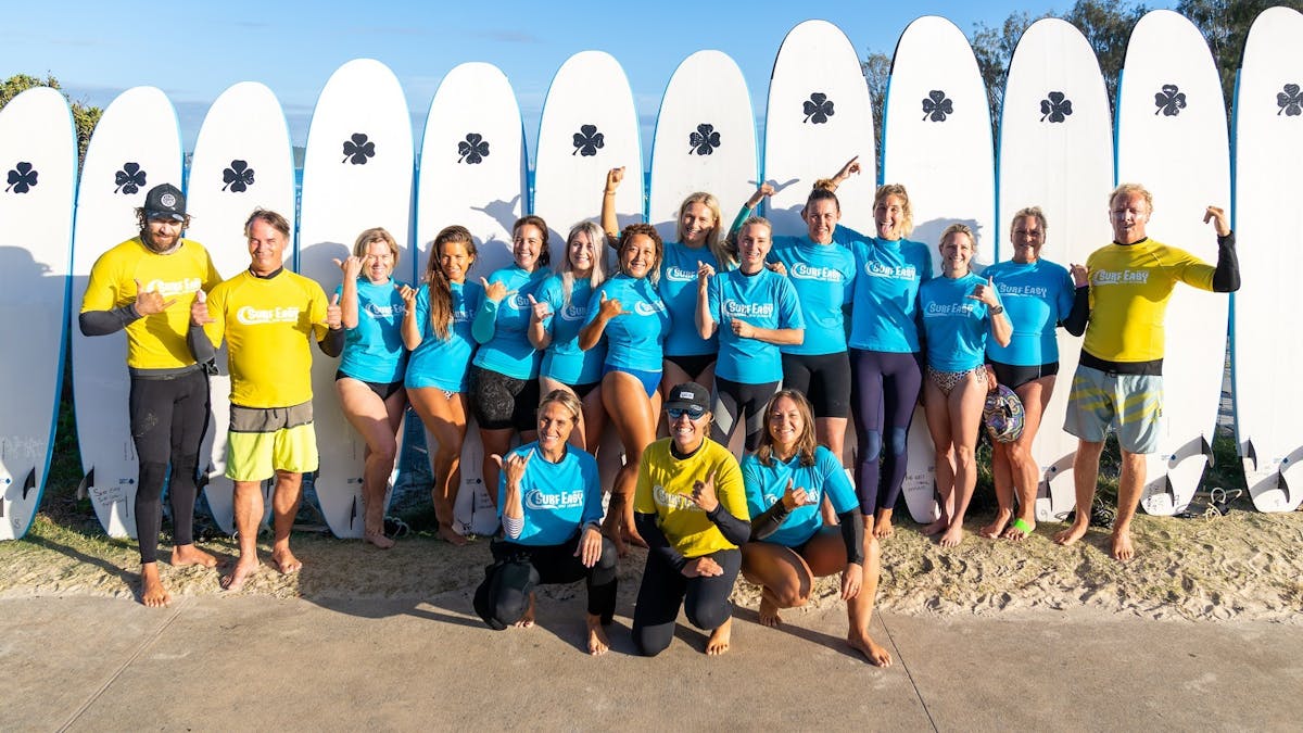Group of ladies and surf instructors with surfboards in back.