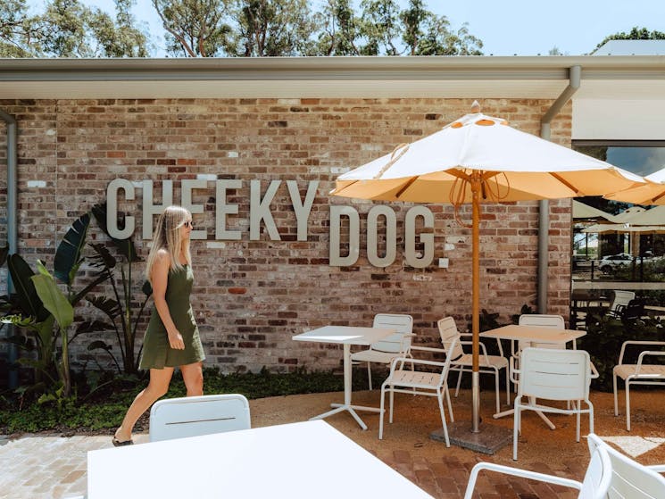 Woman walks between tables, chairs and umbrellas at the entrance of the Cheeky Dog