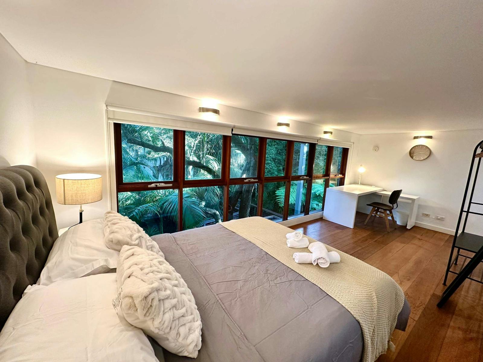 Bedroom featuring a queen-size bed, bedside tables, large windows, and leafy outlook.