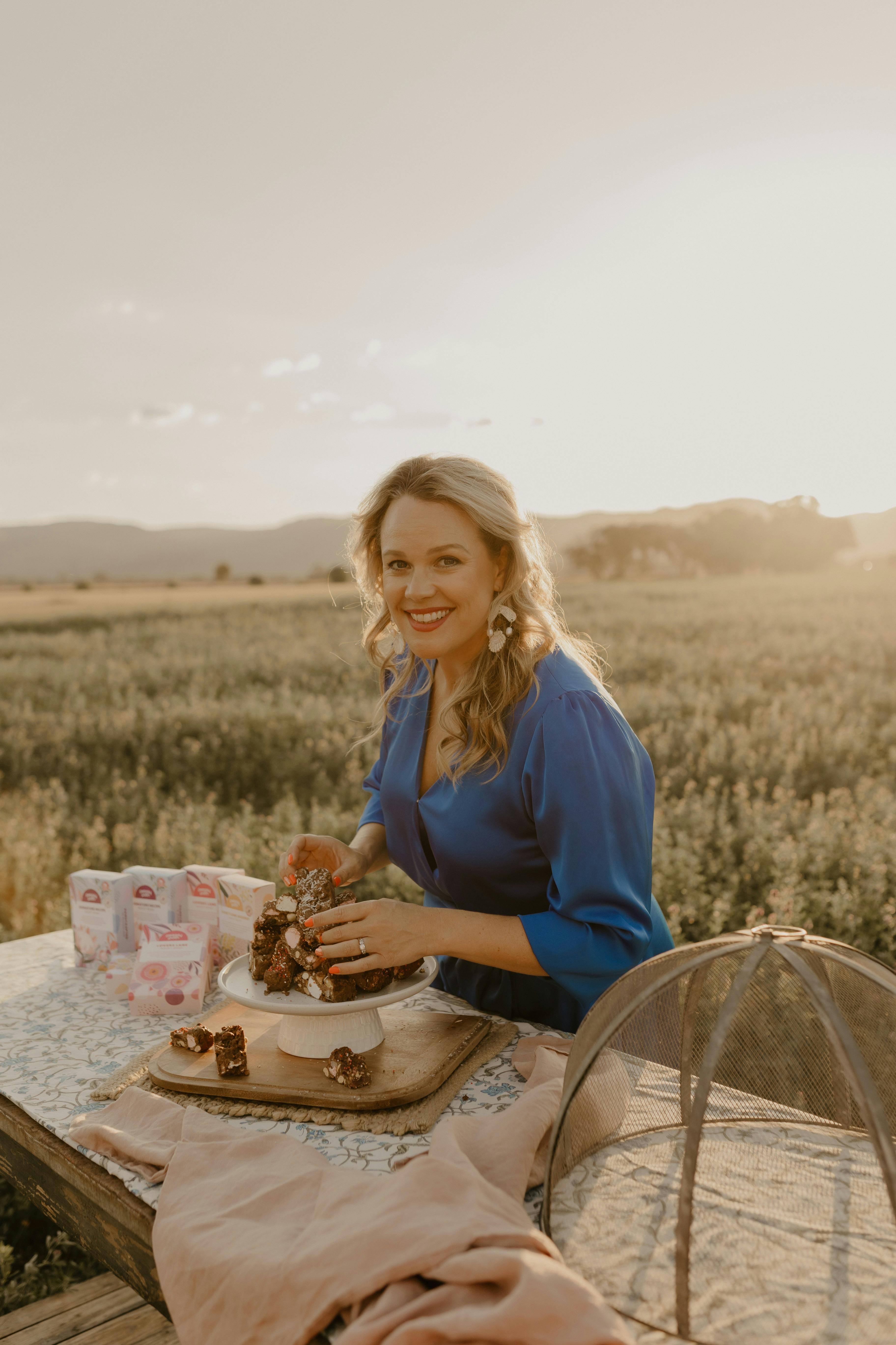Founder in blue dress, in a field of lucerne crop, with a table of rocky road and product boxes