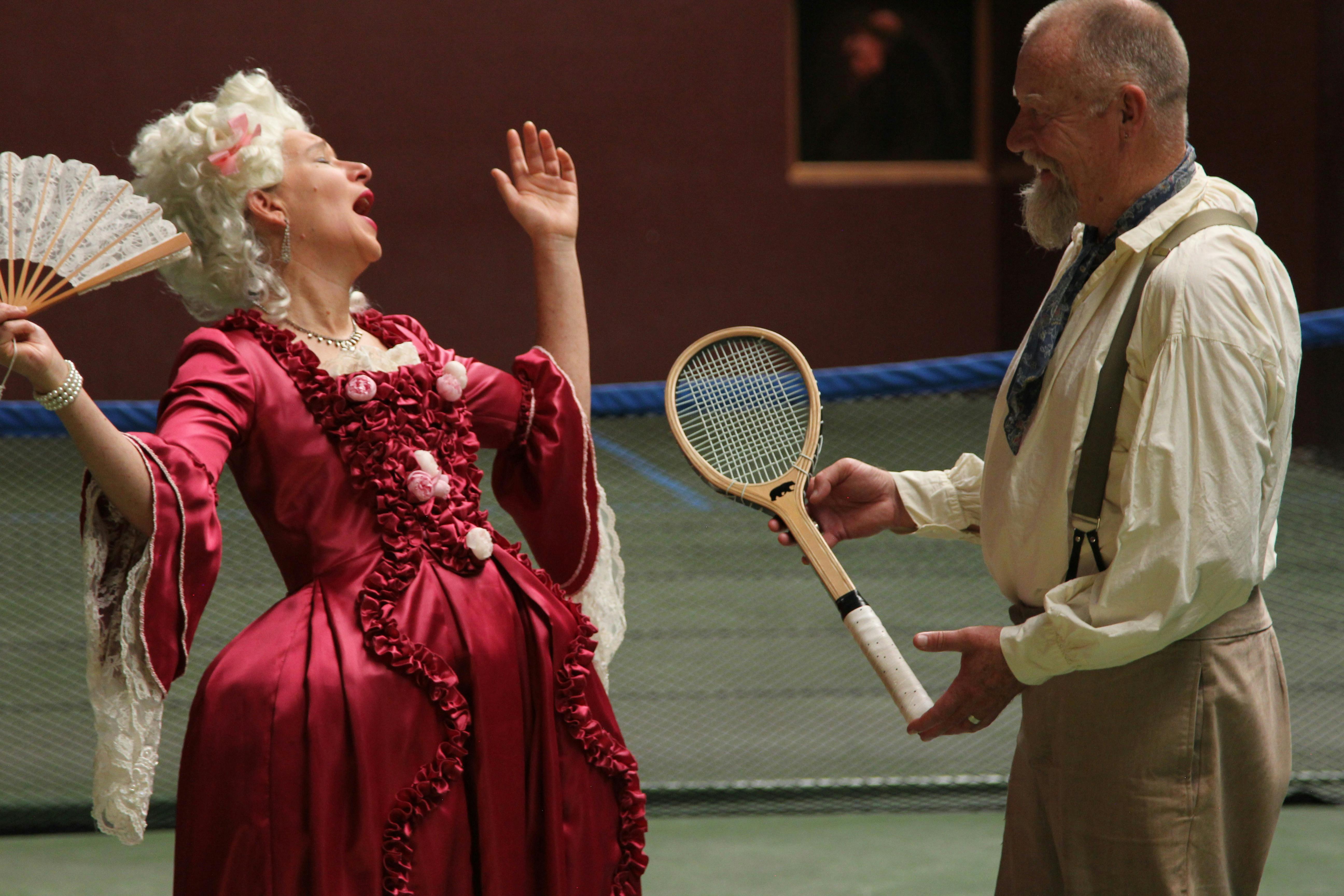 A woman in 18th century dress reacts in surprise to a man in 19th century dress holding a racquet.