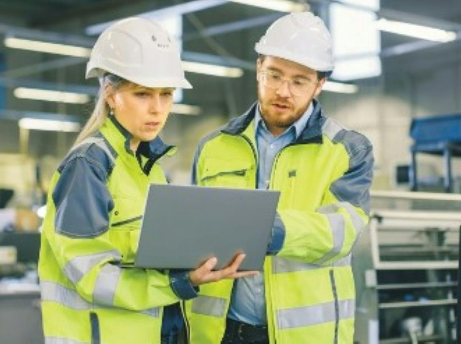 a man and a woman wearing hard hats and hi-vis looking at a laptop that the woman is holding